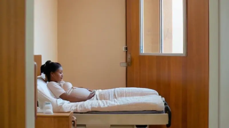 Una mujer embarazada está sentada en una cama hospitalaria gris rodeada por paredes blancas y cubierta con mantas de tono paja